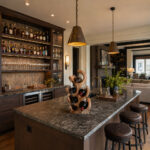 Close-up of the Natural Suar Wood Tree Flow Wine Rack displayed on a dark granite kitchen island, holding bottles, in a sophisticated, rustic modern home interior