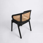 A view from behind the Nara Teak Rattan Wave Chair with a black frame, centered against a white background. The focus is on the large, elegantly curved expanse of the natural rattan weave on the backrest and the minimalist construction of the chair