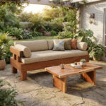 A Chunky Solid Suar Wood Bench with Backrest styled for comfort with a beige linen seat cushion and several patterned (tropical) and plain throw pillows. The solid Suar (Trembesi) wood bench is placed in an outdoor garden patio setting with a matching coffee table, natural stone pavers, lush greenery, and a wooden pergola, creating a relaxing atmosphere